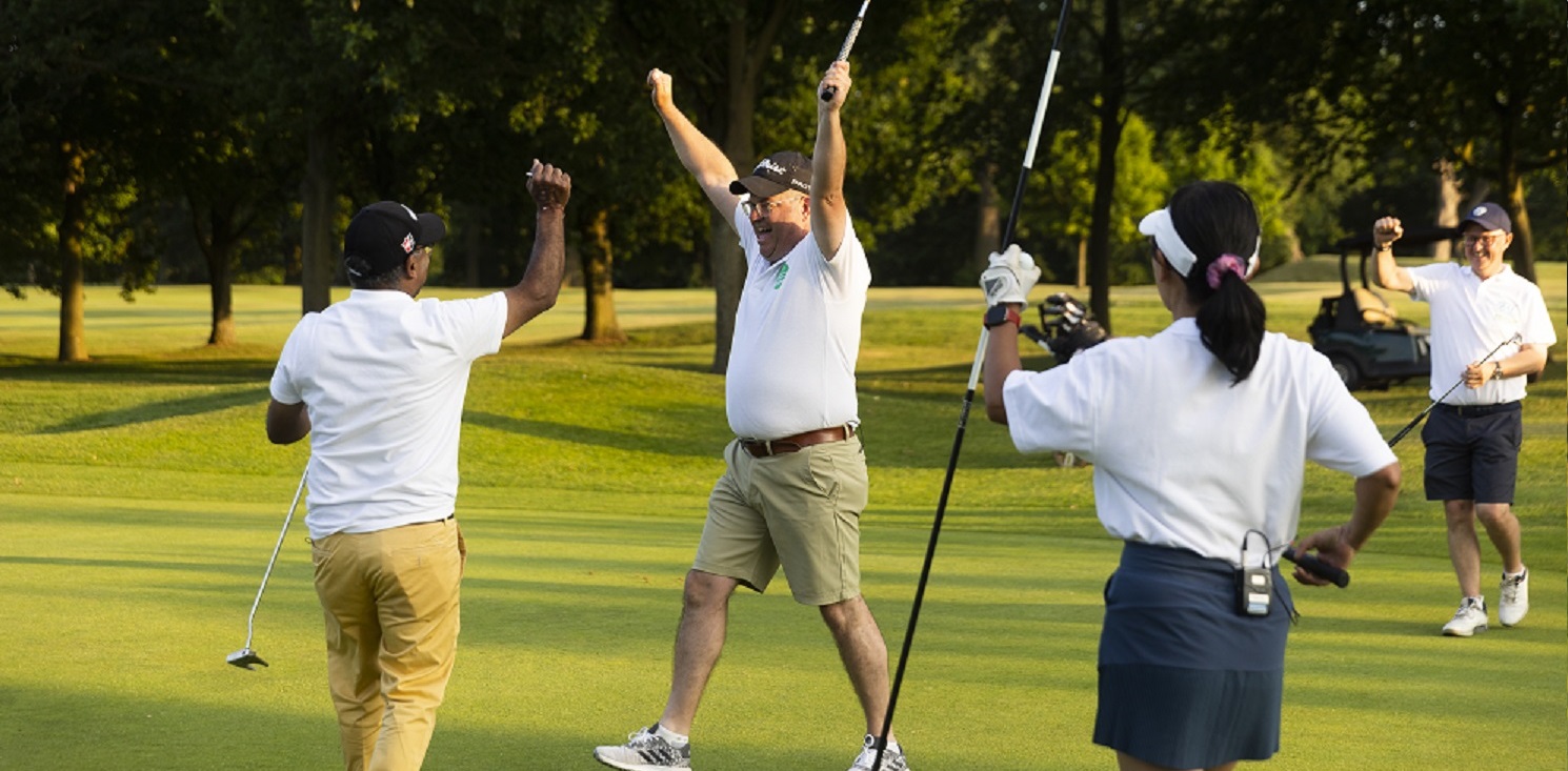 A cheerful group of people on a golf course, with arms raised in celebration, enjoying a sunny day outdoors.