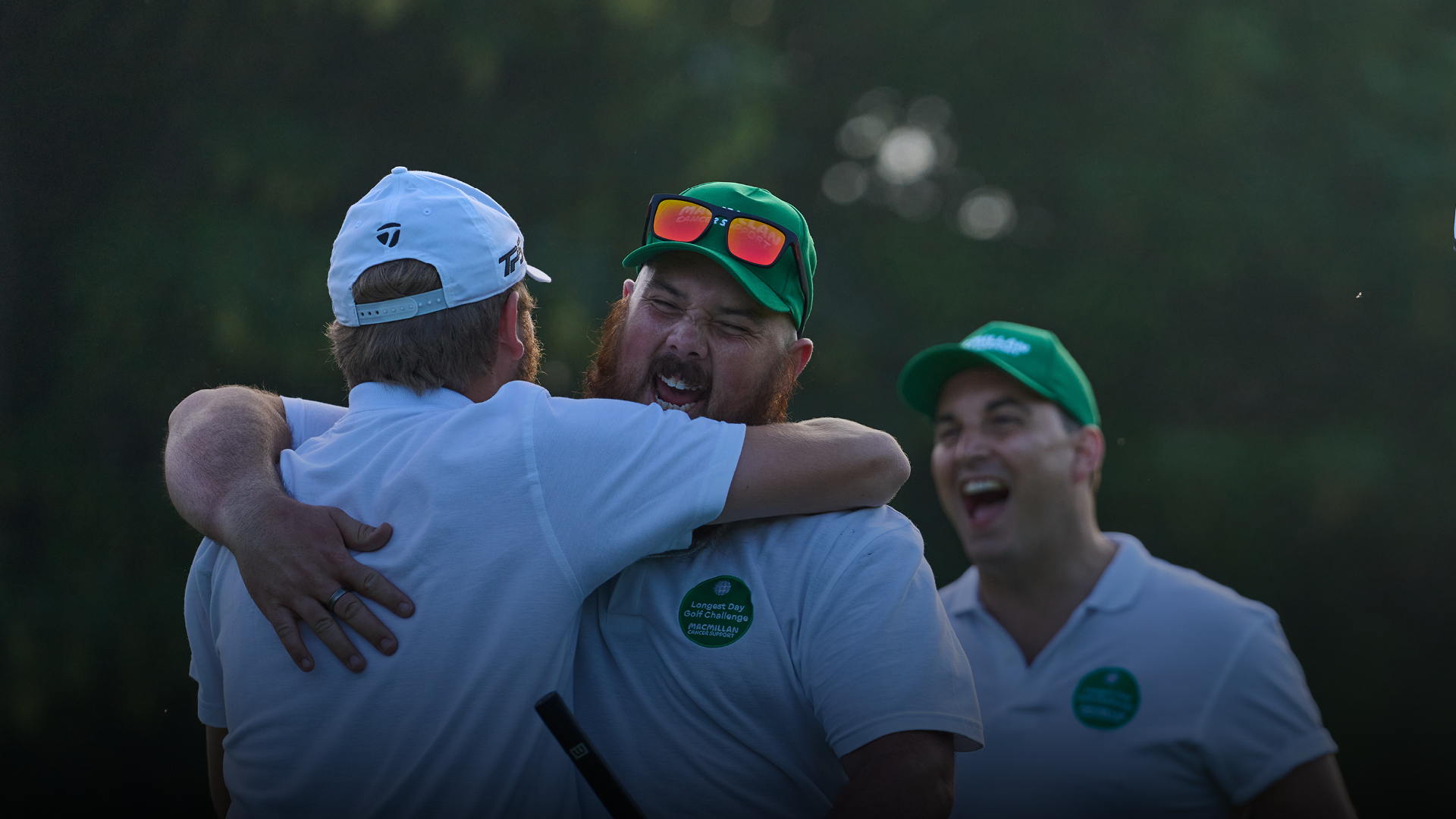 Three men in white shirts and green hats embracing each other with smiles on their faces.