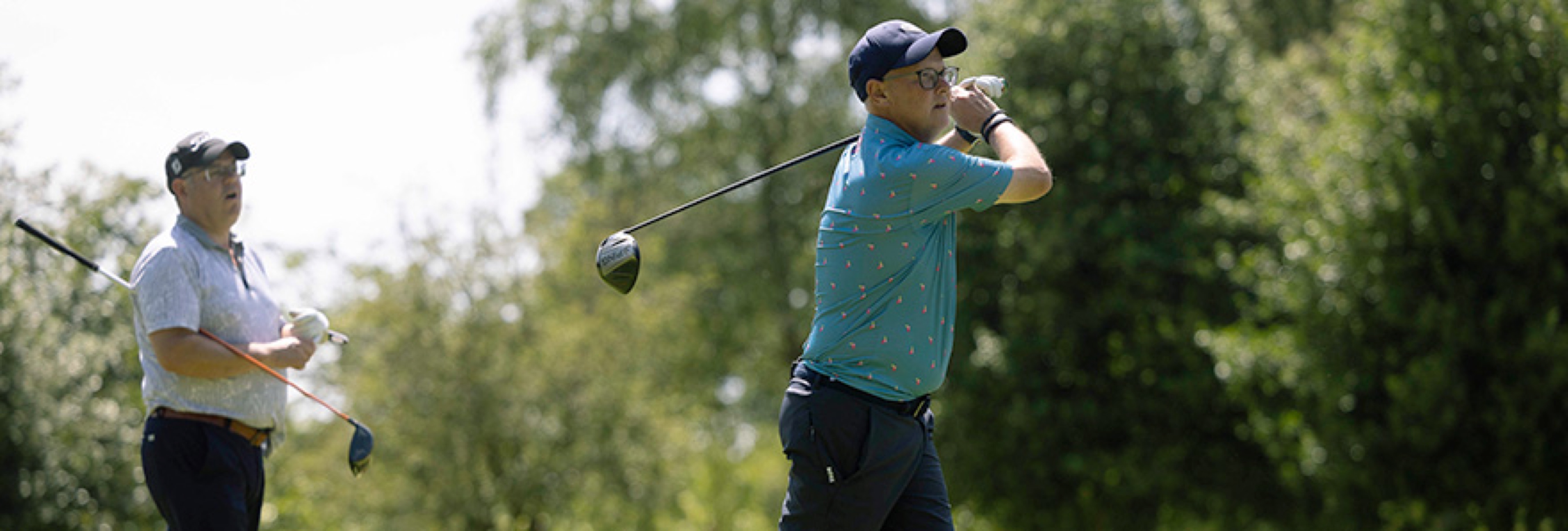 Male golfer taking a shot with another male golfer stood behind him spectating